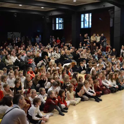 Rodzice i dzieci zgromadzeni w auli szkolnej w czasie świątecznego spotkania dla klas wczesnoszkolnych. 