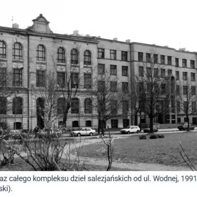 Na zdjęciu widać duży, wielopiętrowy budynek szkoły tworzący część kompleksu salezjańskiego. Budowla ma wiele rzędów okien, a jej lewa część jest bardziej ozdobna architektonicznie. Przed budynkiem znajduje się trawnik, drzewa i zaparkowane samochody. Fotografia została wykonana od strony ulicy Wodnej w 1991 roku.