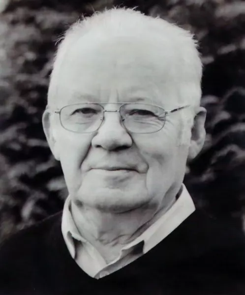 Na zdjęciu widoczny jest ks. Antoni Gabrel. Ma na sobie okulary oraz ciemny sweter z jasną koszulą pod spodem. Patrzy spokojnie w stronę aparatu, a w tle widać rozmyte rośliny.