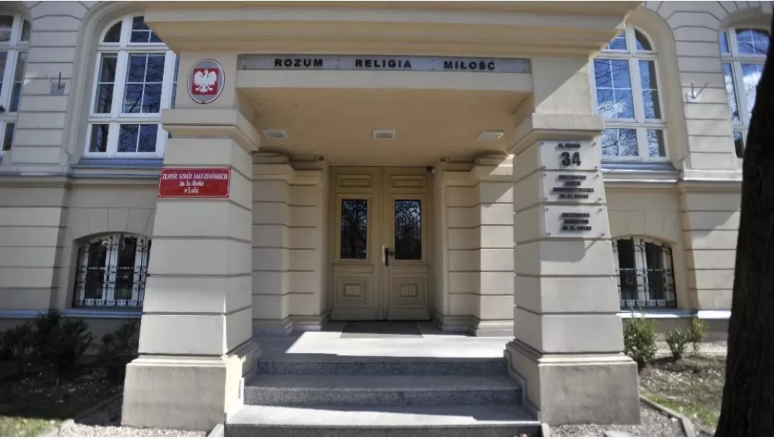 Zdjęcie przedstawia główne wejście do zabytkowego budynku szkoły Zespołu Szkół Salezjańskich w Łodzi im. ks. Bosko, z jasną elewacją i symetryczną architekturą. Nad drzwiami widnieje hasło „Rozum – Religia – Miłość”, a przy wejściu umieszczone są tablice informacyjne oraz godło państwowe.